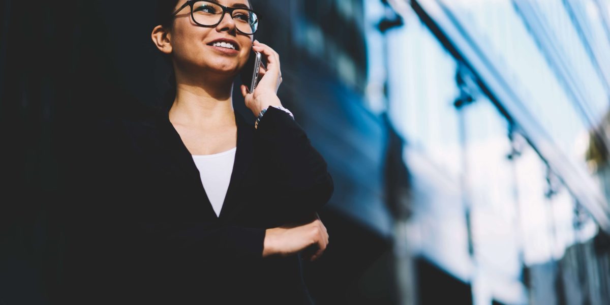 Prosperous businesswoman in eyeglasses and stylish black suit smiling during mobile conversation on modern smartphone standing outdoors near office building.Copy space area for financial information